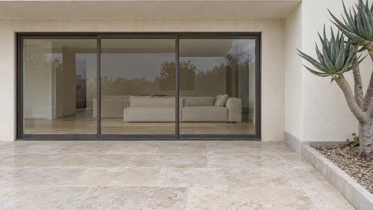 Ivory stucco home opening onto a limestone terrace through closed bronze-framed glass doors, with a sculptural branching aloe in a curb-edged planting bed.
