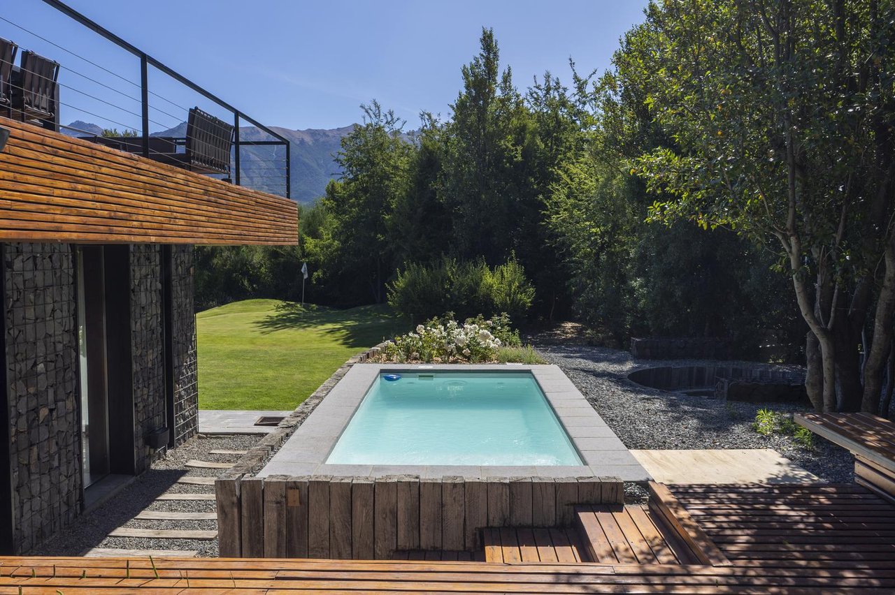 Outdoor pool beside a modern stone-and-timber home, with a timber deck and mountains beyond the garden.