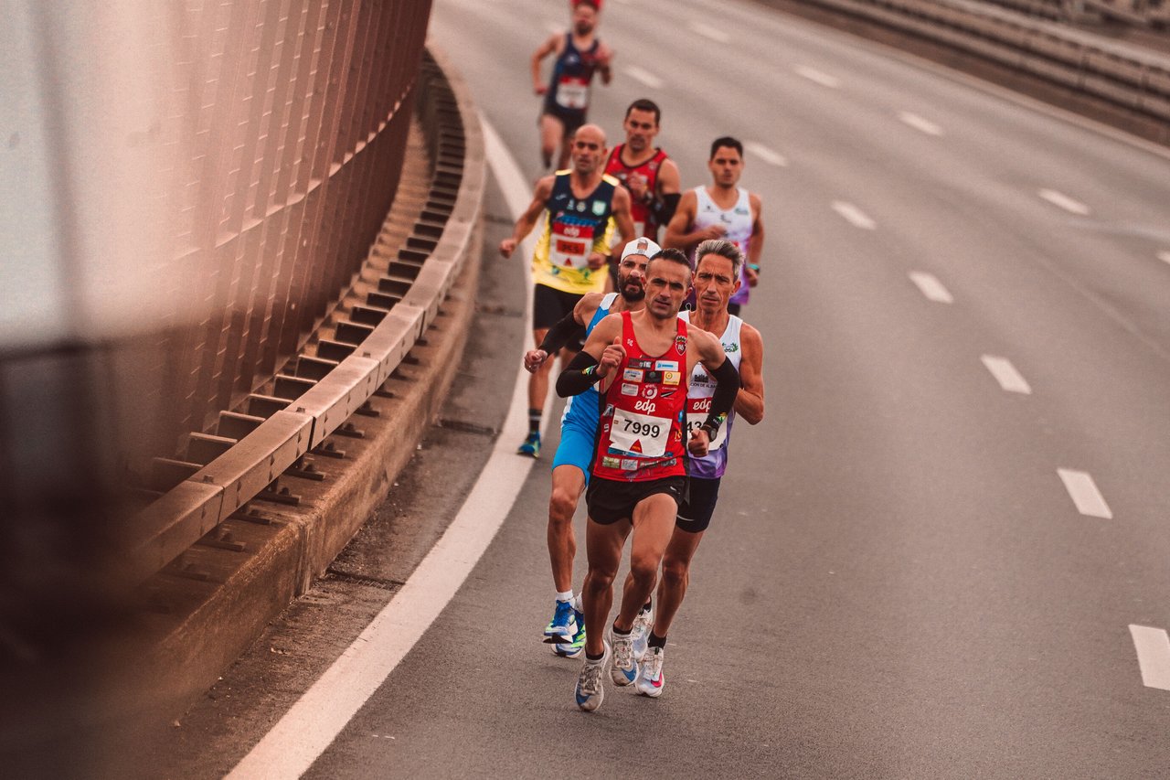 A panoramic photograph of thousands of runners crossing the Verrazzano-Narrows Bridge during the New York City Marathon, with the New York Harbor and Brooklyn skyline in the background.