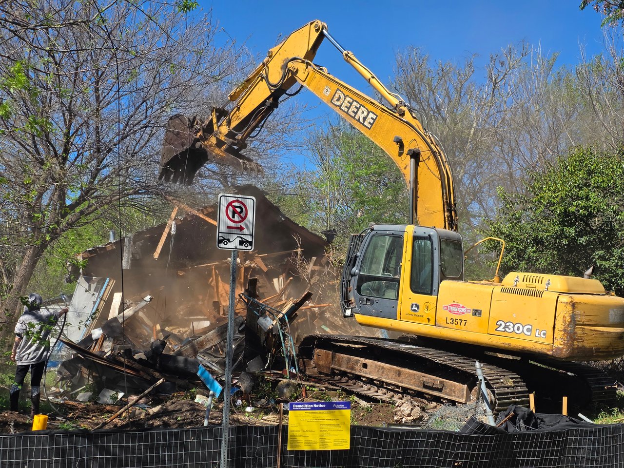 An excavator demolishes a house amid rubble at a fenced development site.