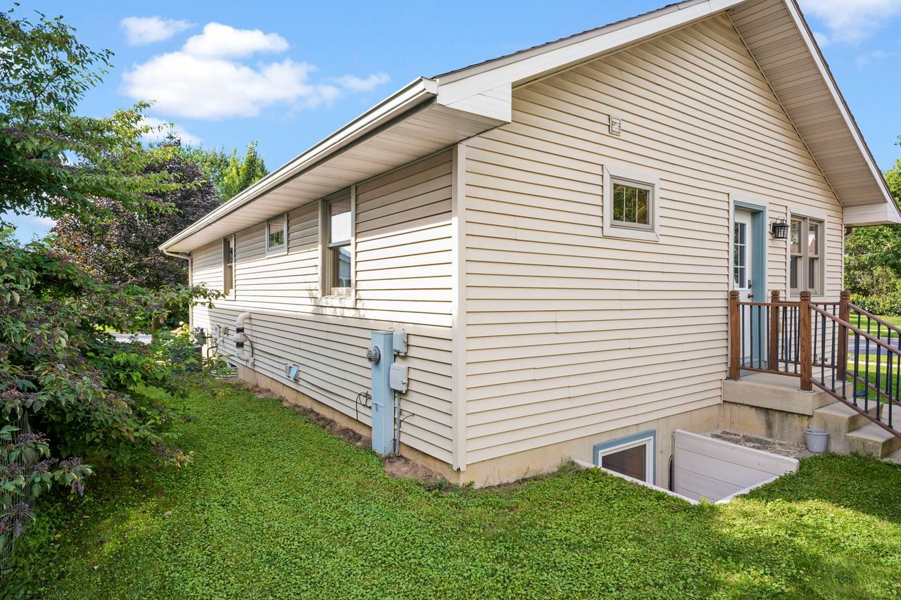 The side of a Madison-area home where the siding meets the exposed foundation, with a basement window well at grade and lawn sloping along the wall.