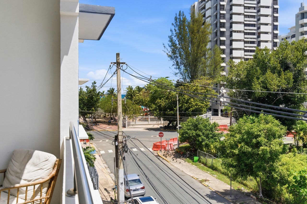 Apartment balcony overlooking a tree-lined street and modern high-rises, with a wicker chair in the foreground.