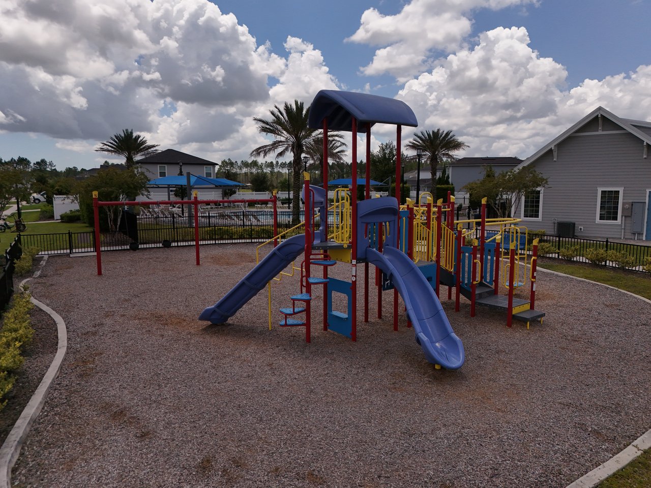 Community playground with blue slides and yellow climbing equipment behind a black fence, surrounded by homes and palm trees.