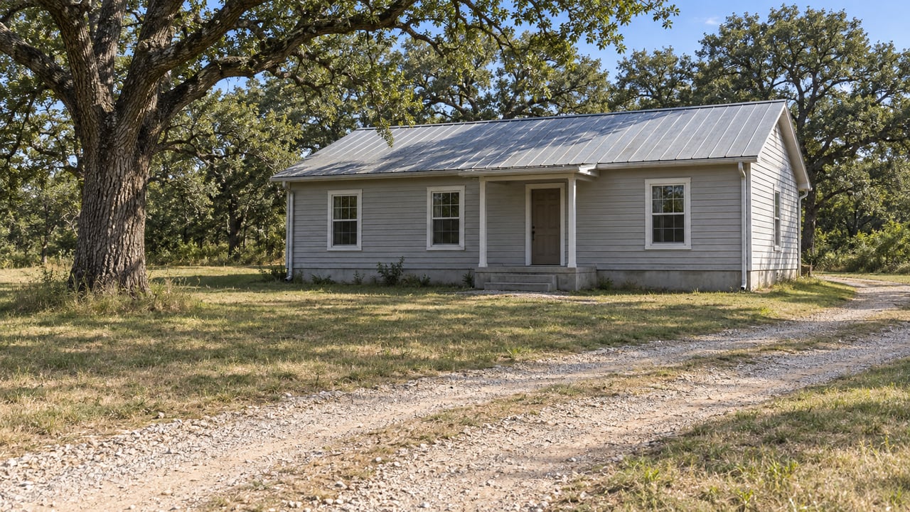 A modest gray wood-sided home with a metal roof sits beyond a broad grassy clearing and pale gravel drive beneath mature post oaks.