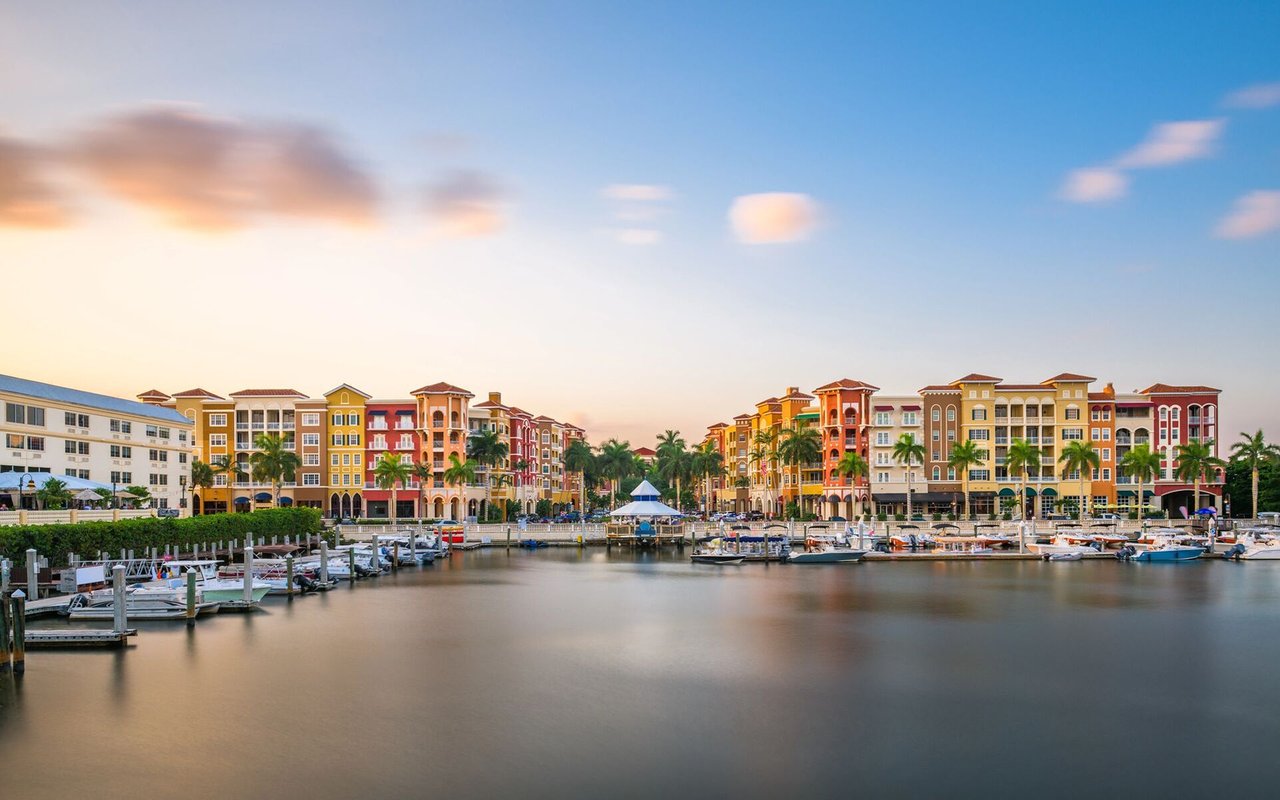 Waterfront marina lined with colorful buildings, palm trees, and docked boats at sunset.