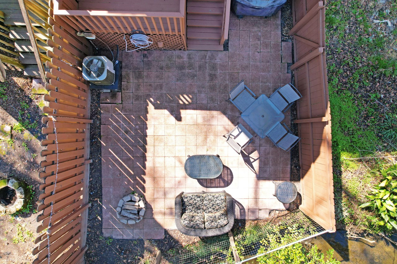 Aerial view of a fenced patio with a square table, four chairs, wicker loveseat, small table, and fire pit. Surrounded by greenery and shadows.