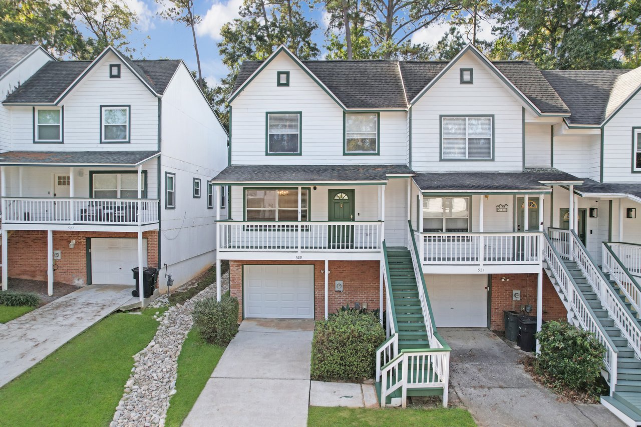 Front exterior view of the townhome at 529 Collinsford Road, Tallahassee, Florida, showing a two-story design with white siding, green trim, and a one-car garage. The home features a covered front porch with white railings, a green front door, and a green staircase leading up to the main entrance.