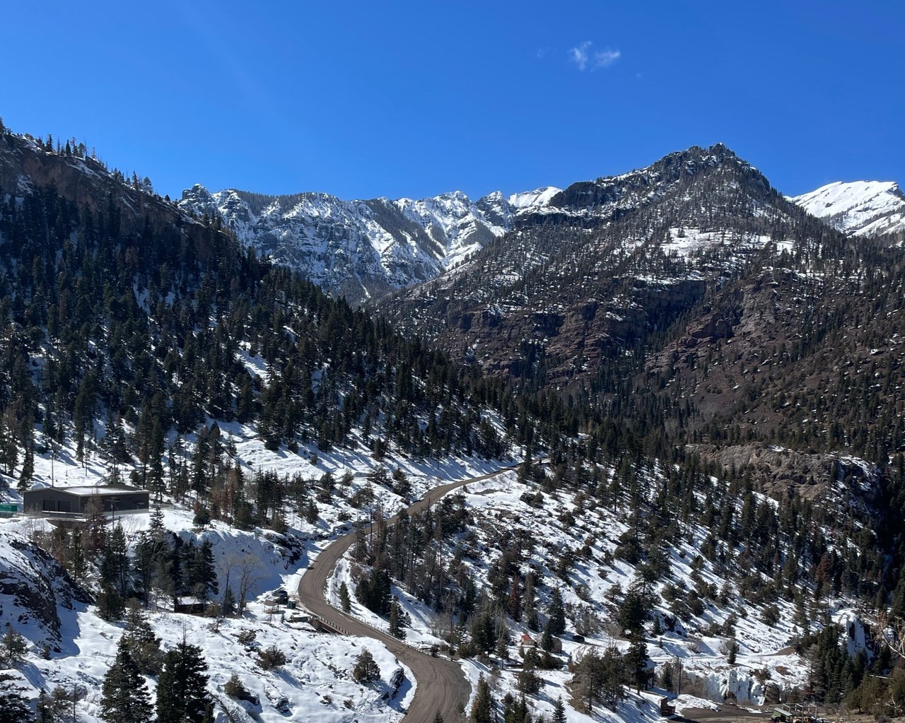 Winding road through a snowy mountain valley, surrounded by evergreen forest and rugged peaks.