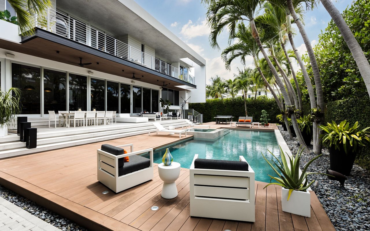Modern home beside a turquoise pool, with a wood deck, white lounge furniture, and tall palms.