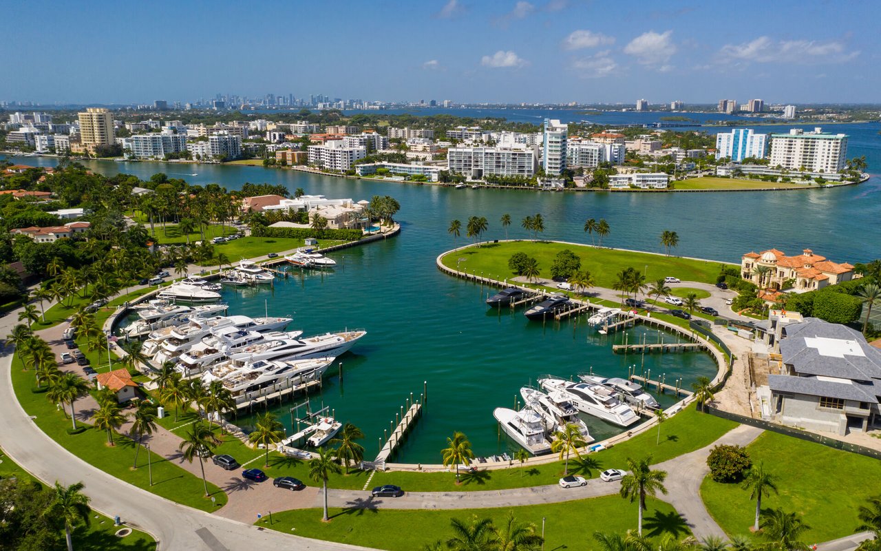 Aerial view of a waterfront neighborhood with yacht-filled marinas, palm-lined lawns, and a distant city skyline.