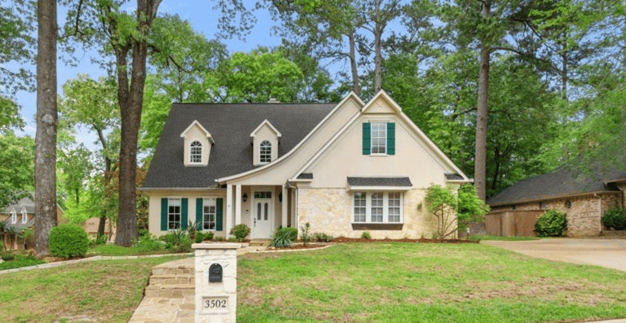 Front exterior of a cottage-style home with cream stucco and stone facade, dark roof, green shutters, dormers, and mature pine and oak trees in The Woods neighborhood, Tyler, TX