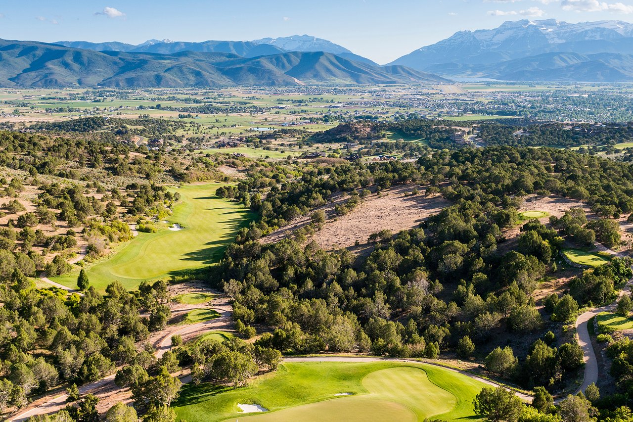 Aerial view of a golf course winding through wooded hills, with a valley and mountains beyond.