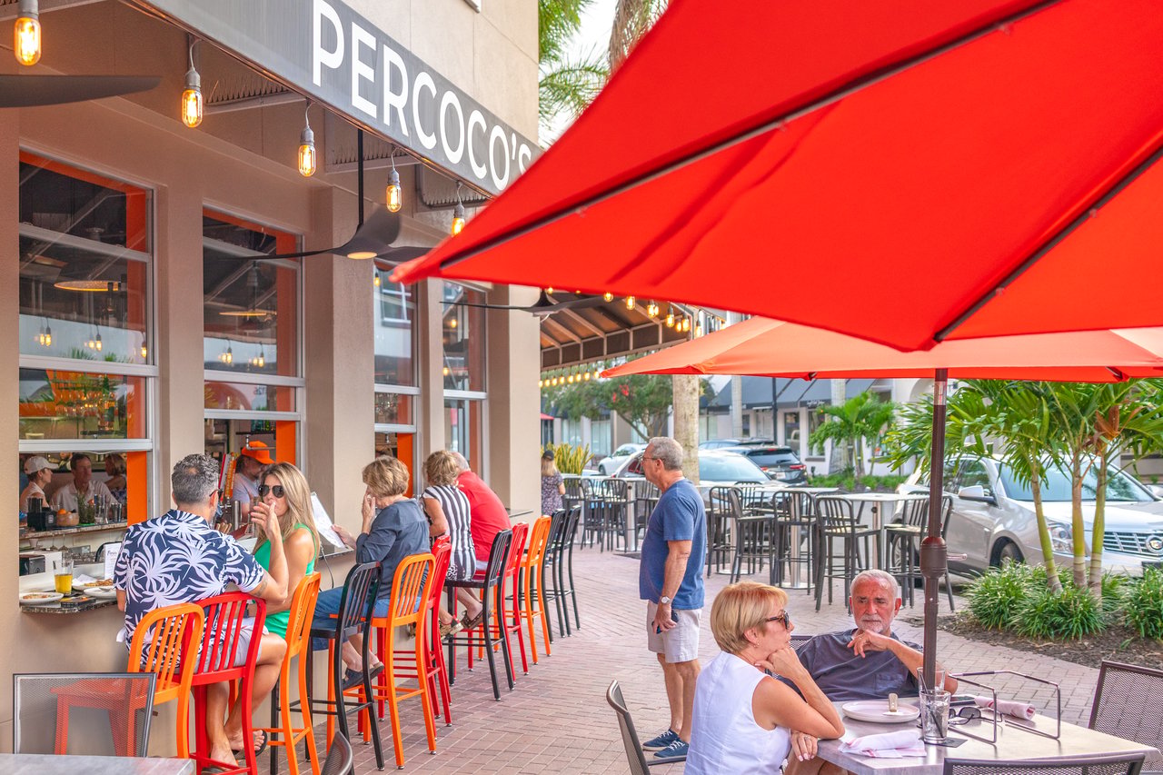 Restaurant patio with diners beneath bright red umbrellas beside the PERCOCO'S storefront.