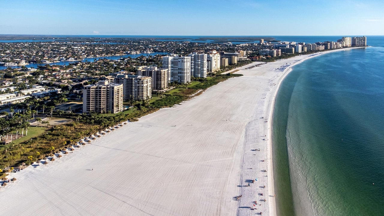 Aerial view of a long white-sand beach, Gulf water, and coastal condominium towers.