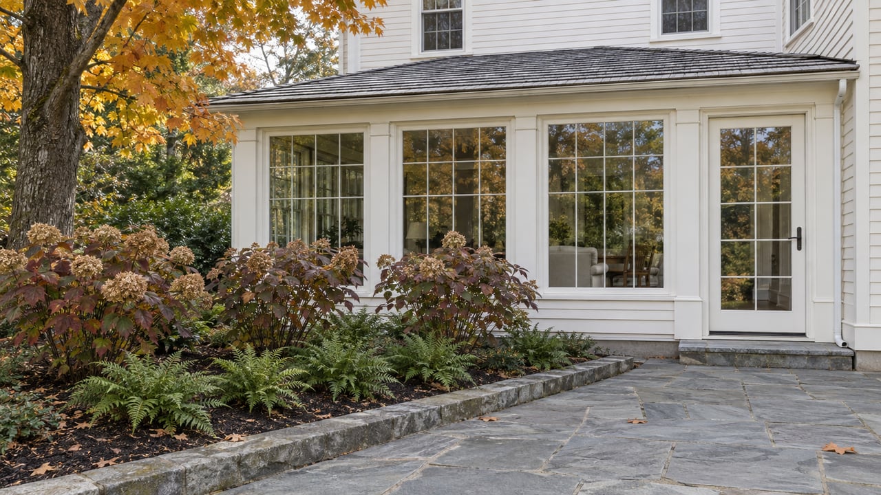 Ivory clapboard home with an enclosed sunroom, bluestone terrace, and a garden of autumn maple, oakleaf hydrangeas, and evergreen ferns.