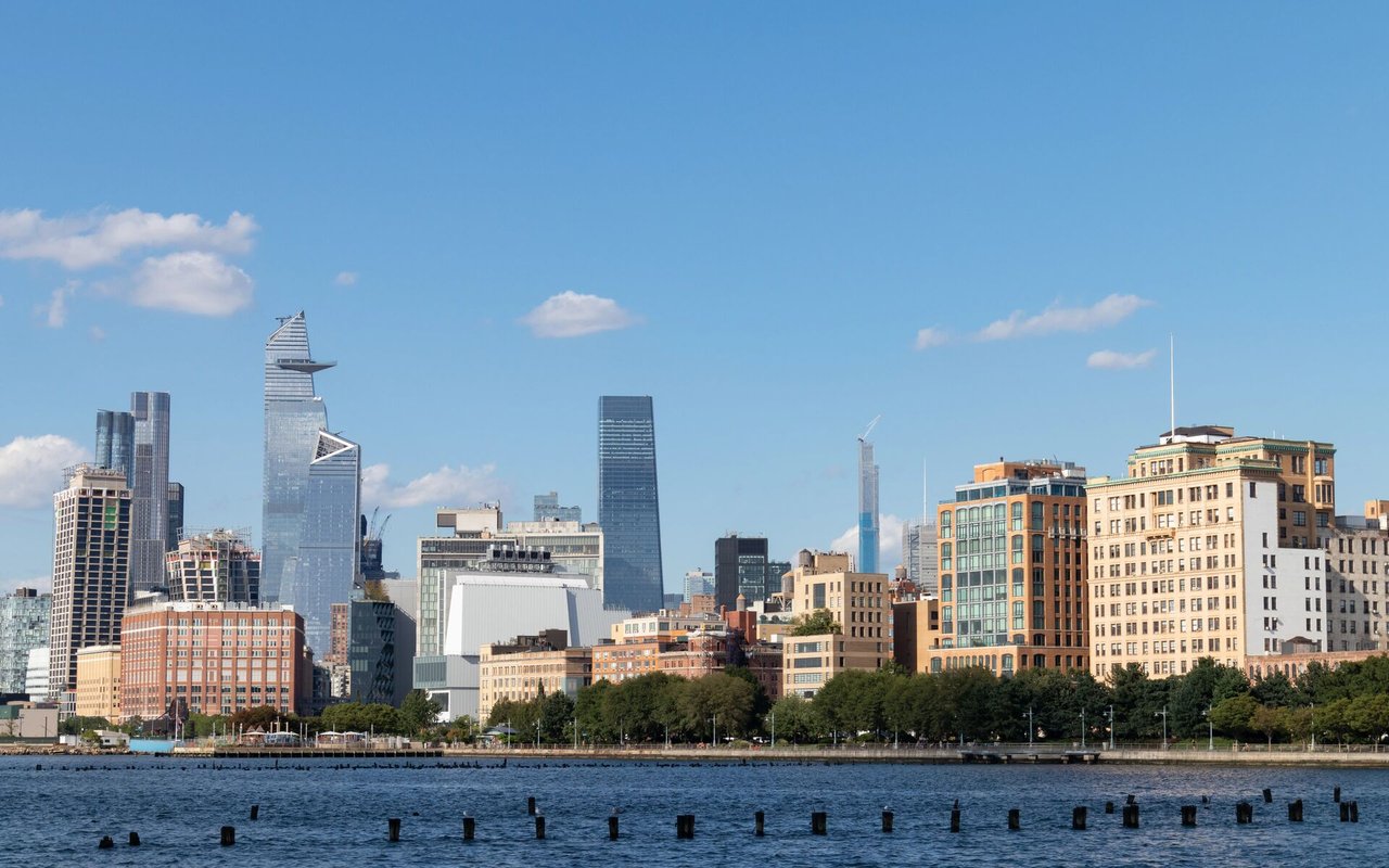 Hudson Yards skyline across the water, with modern towers and a tree-lined waterfront.