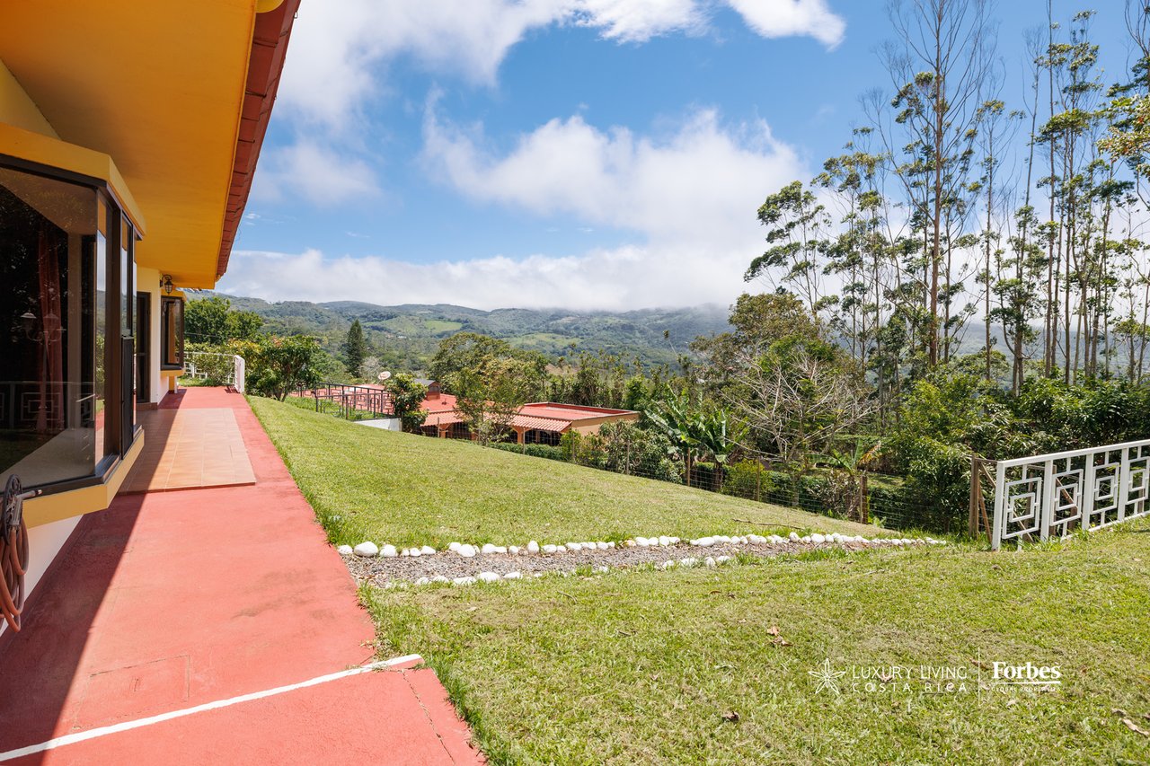 Mountain home beside a red terrace and lawn, overlooking green ridges beneath a cloudy sky.