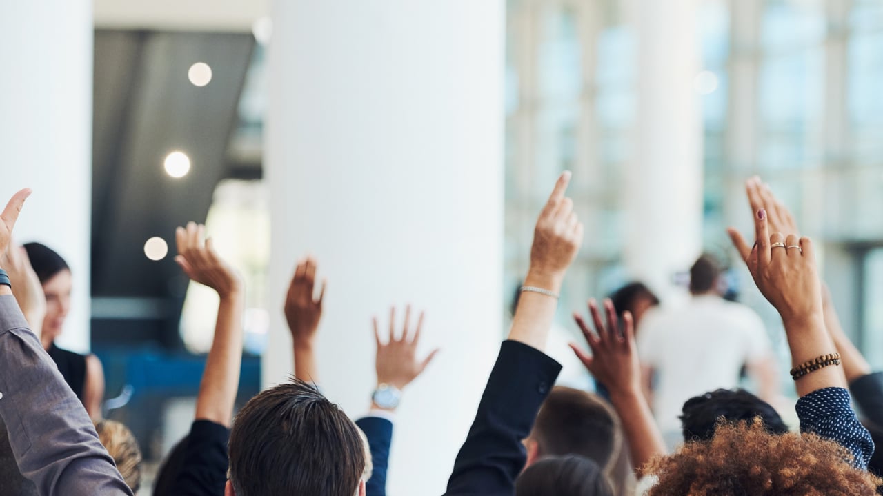 Meeting attendees raise their hands during a discussion in a bright room.