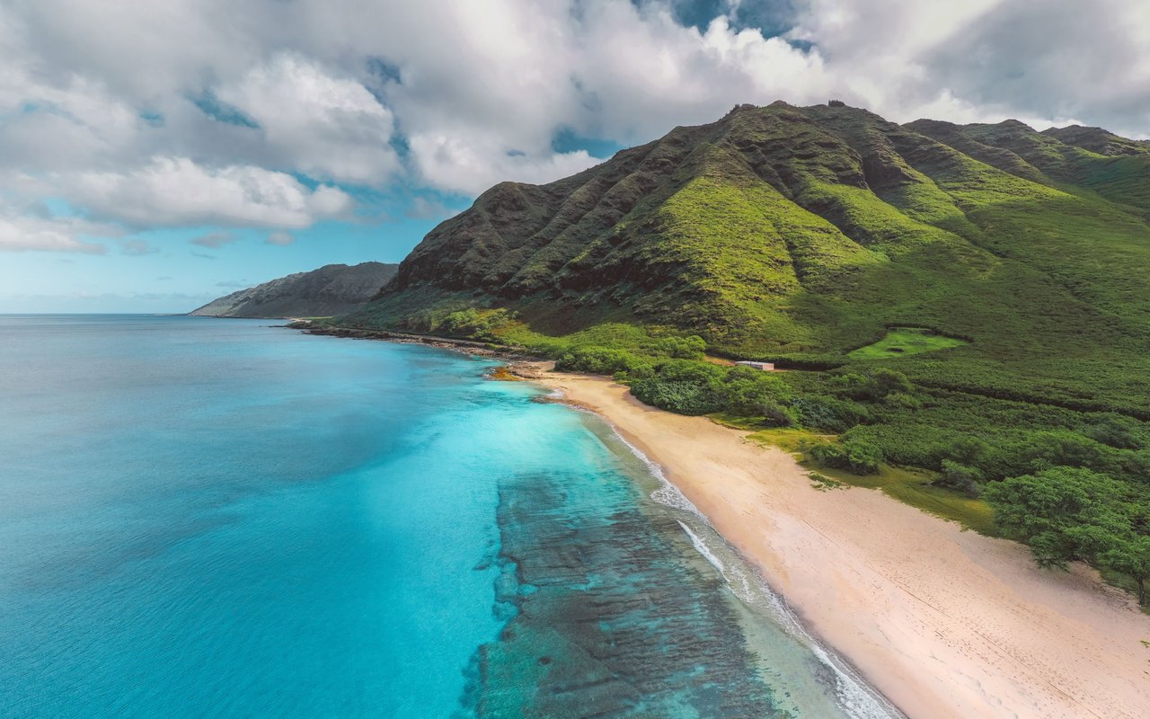 Turquoise water and a sandy beach curve beneath steep, green mountain ridges.