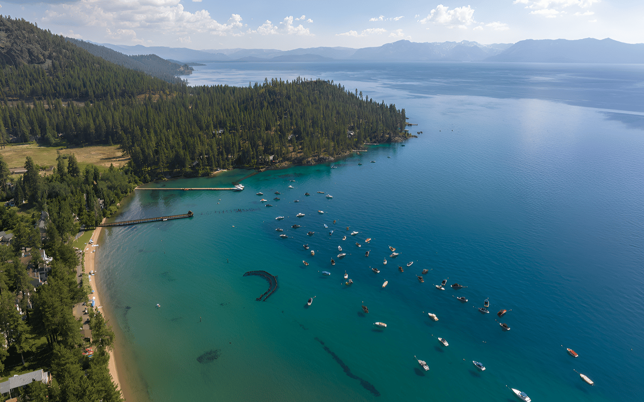 Aerial view of Lake Tahoe’s forested shore, turquoise water, beach, and scattered boats.