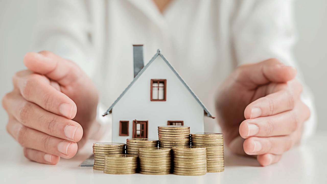 Hands surround a model house and stacks of coins, illustrating saving to buy a home.