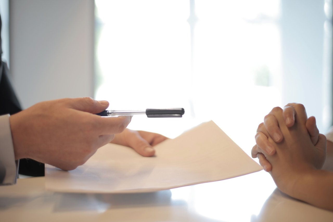 One person offers a pen beside a document as another clasps their hands.