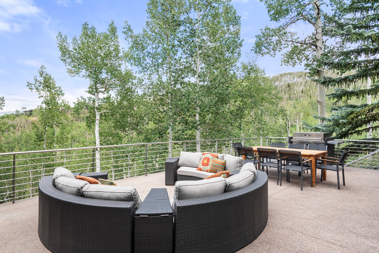 Outdoor patio with curved wicker seating and dining area overlooking a wooded hillside.