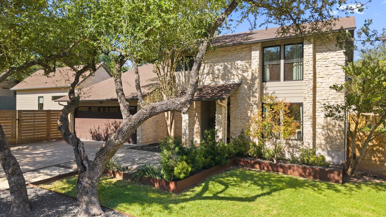 Stone two-story home with a brown garage door, landscaped lawn, and mature trees in front.