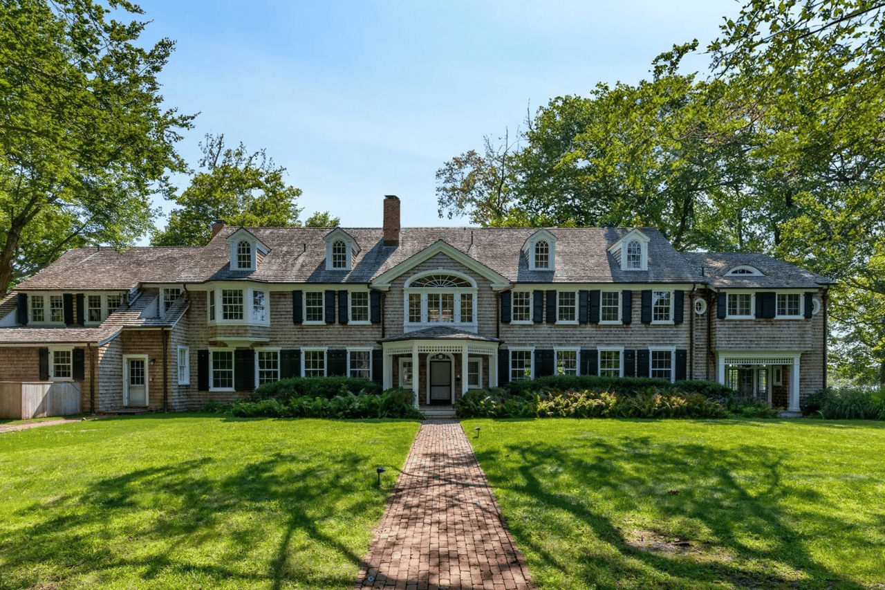 Large shingle-style house with dormer windows, dark shutters, and a brick walkway across the lawn.