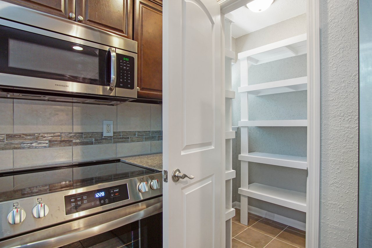 Modern kitchen corner with stainless steel microwave above an oven, next to an open door revealing a well-lit pantry with white shelves.