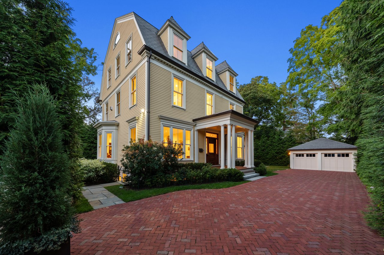 Victorian house with glowing windows, columned porch, detached garage, and brick driveway at dusk.
