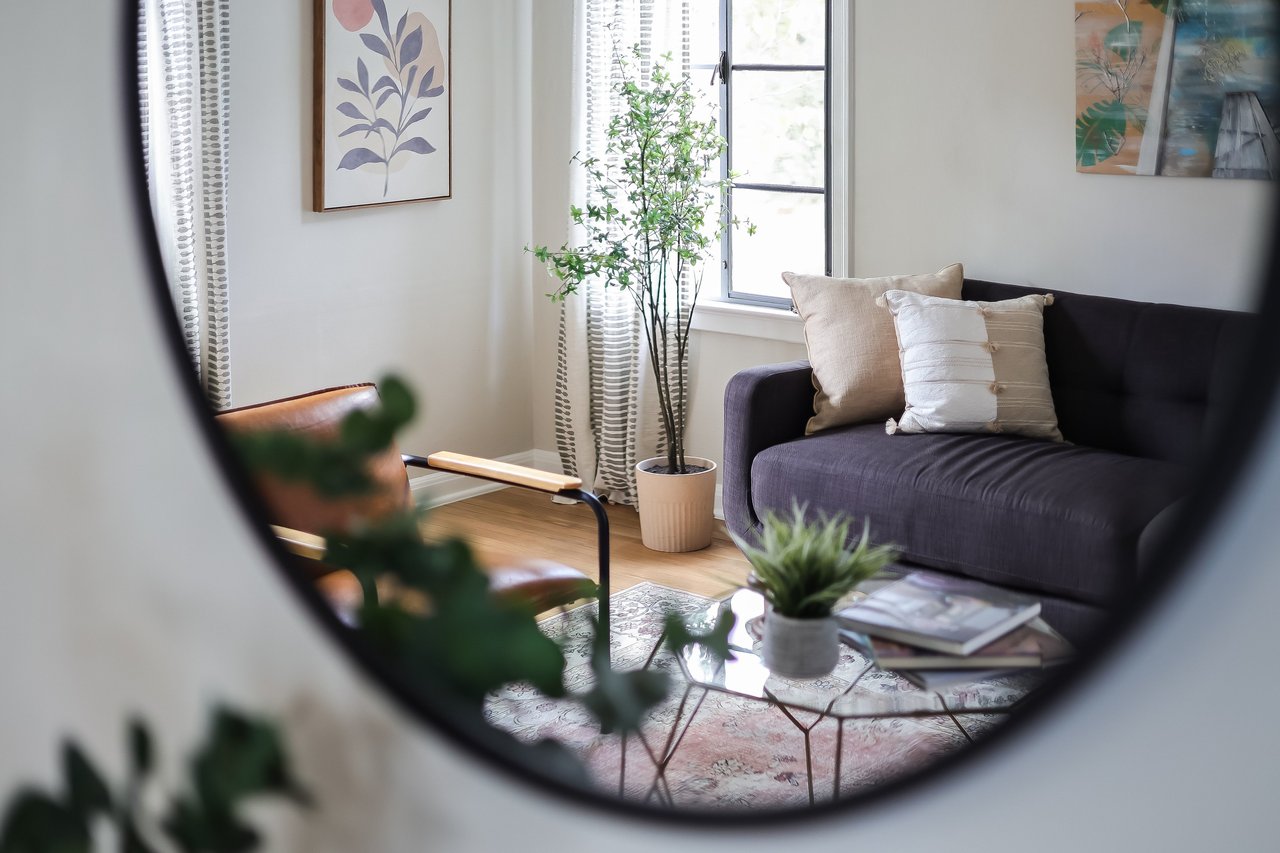 Living room with a dark sofa, wood-and-leather chair, and leafy plants, seen through a round mirror.