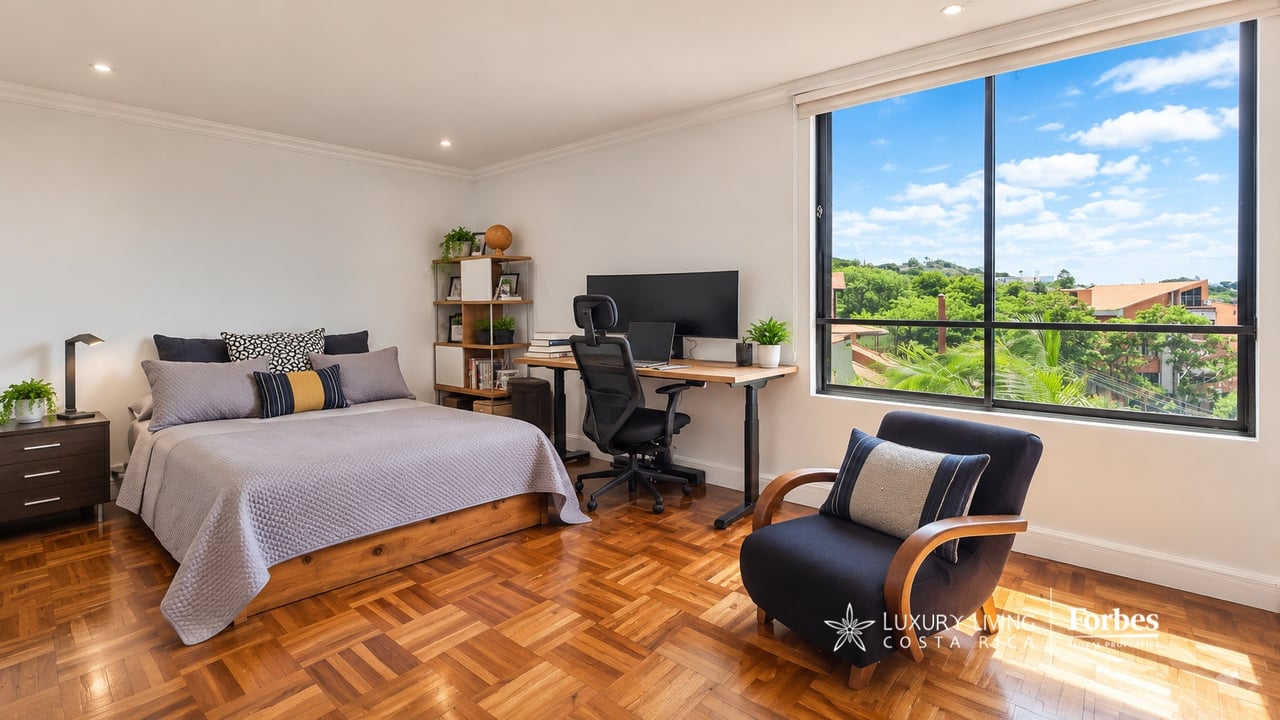 Furnished bedroom with a desk and armchair beside a large window overlooking trees and nearby buildings.