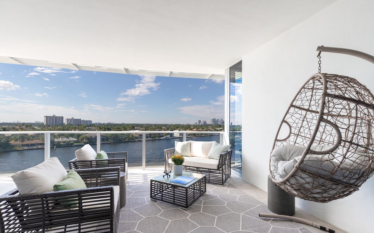 Waterfront terrace with wicker seating and a hanging egg chair overlooking the water and distant skyline.