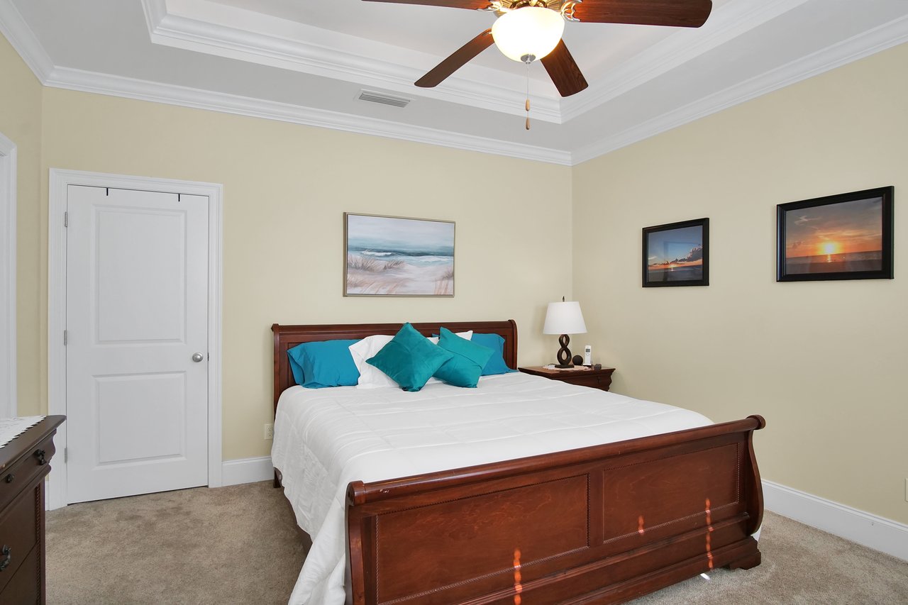 Cozy bedroom featuring a wooden bed, white bedding, and teal pillows. Art on walls, ceiling fan, and soft lighting create a serene, inviting atmosphere.