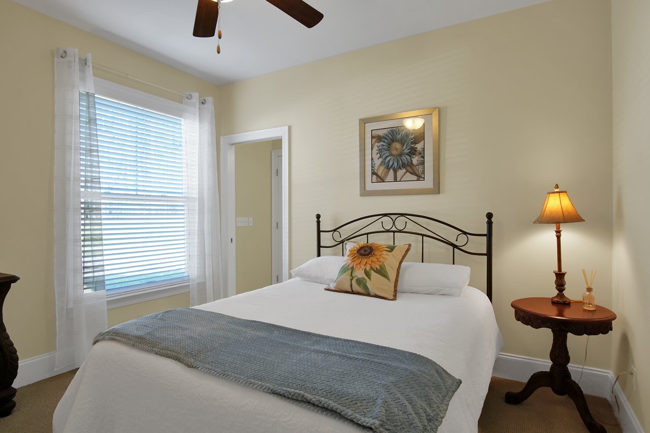 Cozy bedroom with soft yellow walls, featuring a white bed with a sunflower pillow and gray throw. A framed flower picture, wooden side table, and warm lamp enhance the inviting feel.