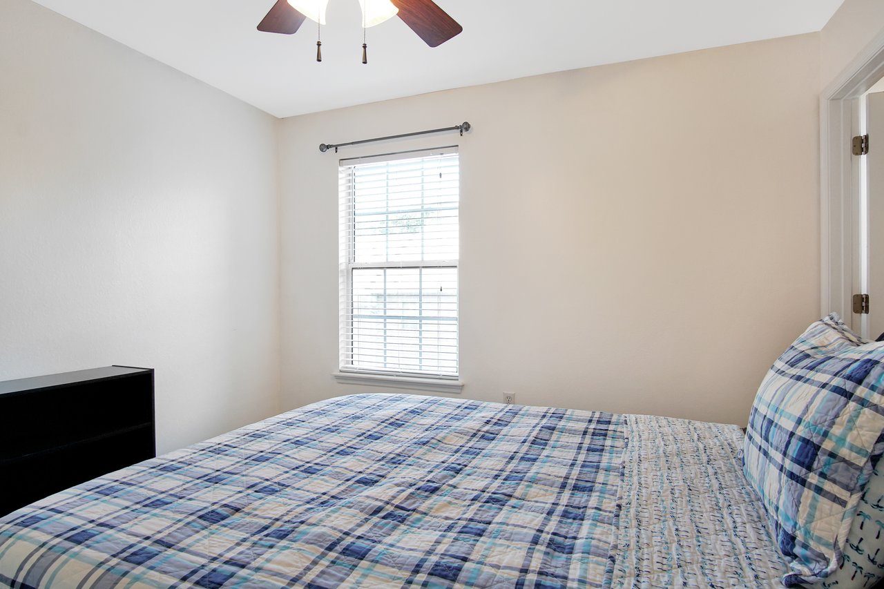 Small bedroom with light walls, a window with blinds, and a ceiling fan. A bed with blue and white plaid bedding adds a cozy touch. Minimalist, serene space.