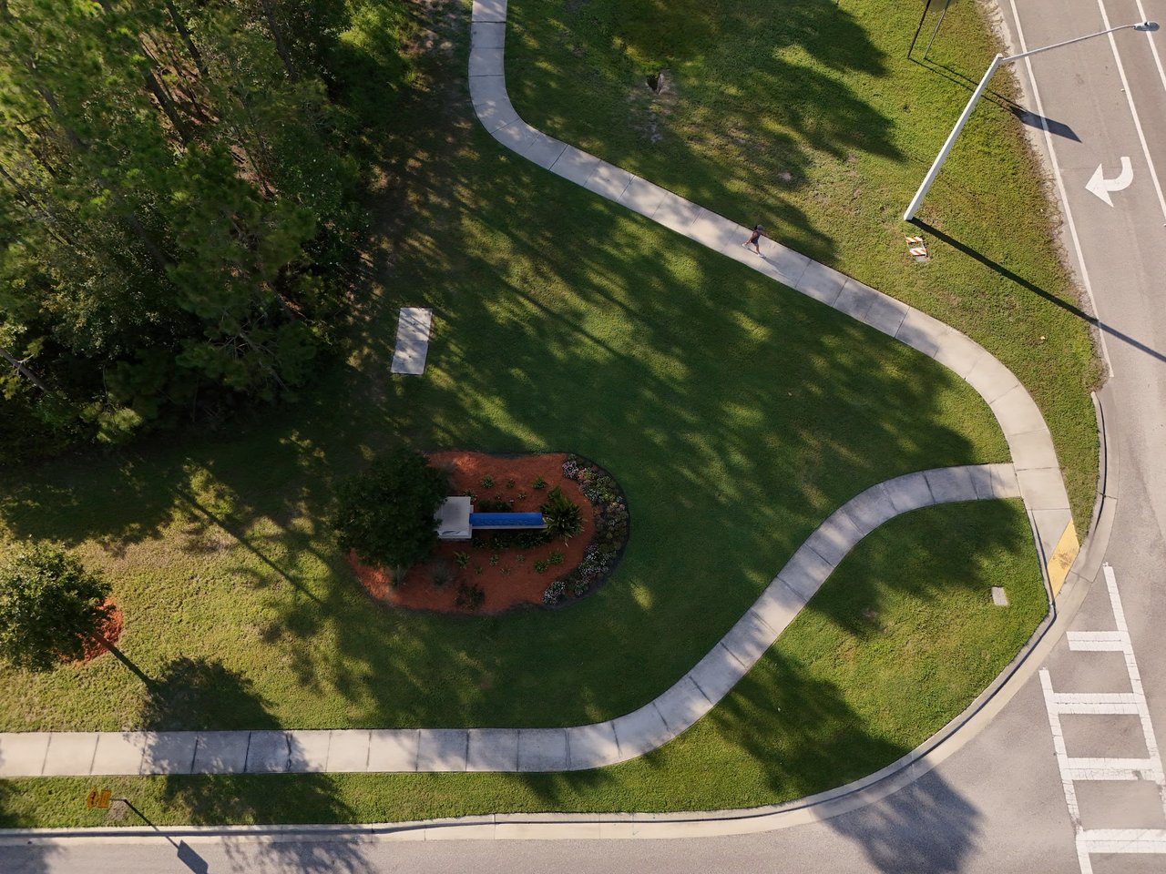 Aerial view of a landscaped community corner with winding sidewalks, lawn, trees, and a pedestrian.