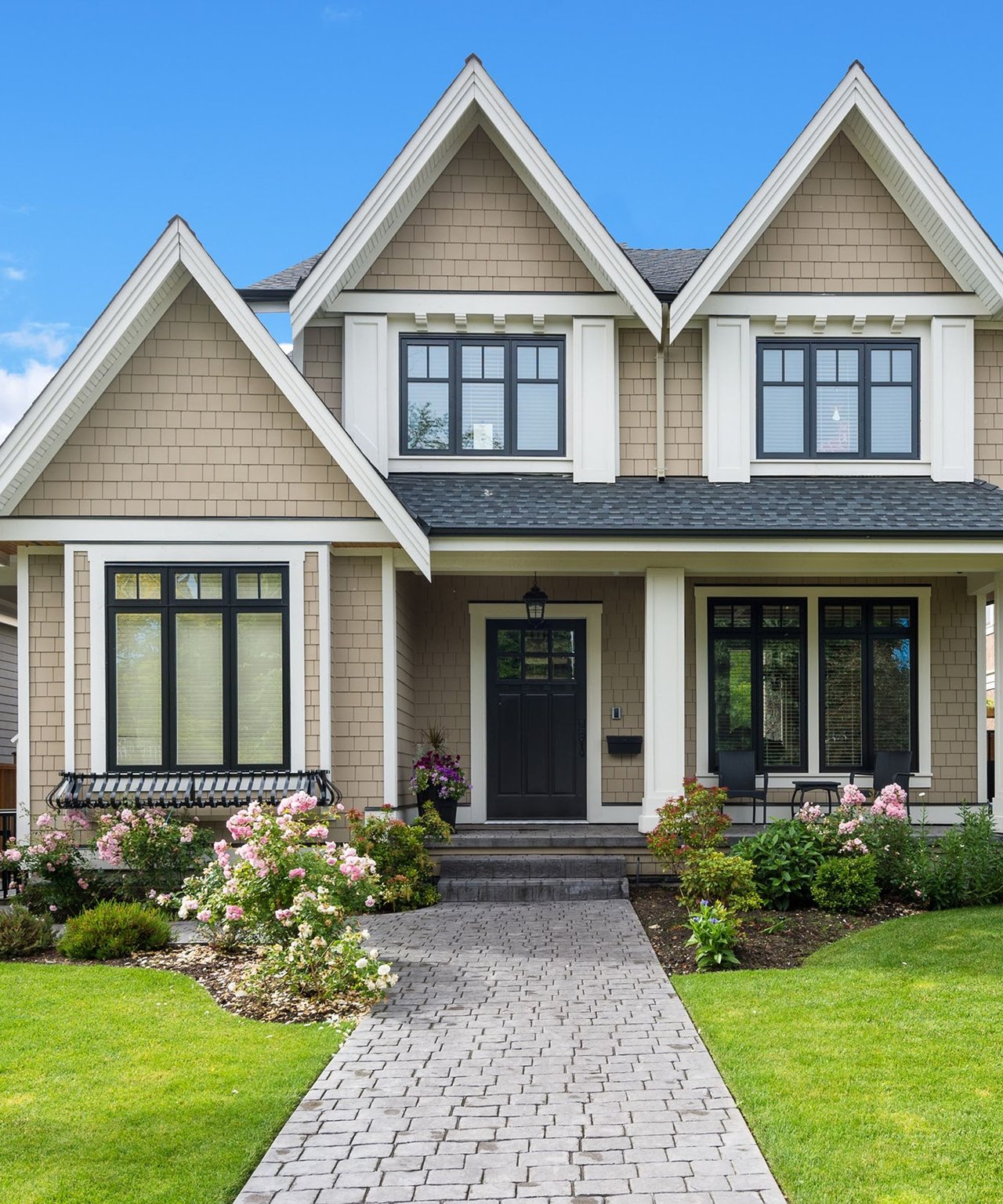 Two-story beige home with three gables, a stone walkway, and pink-flowered garden beds.