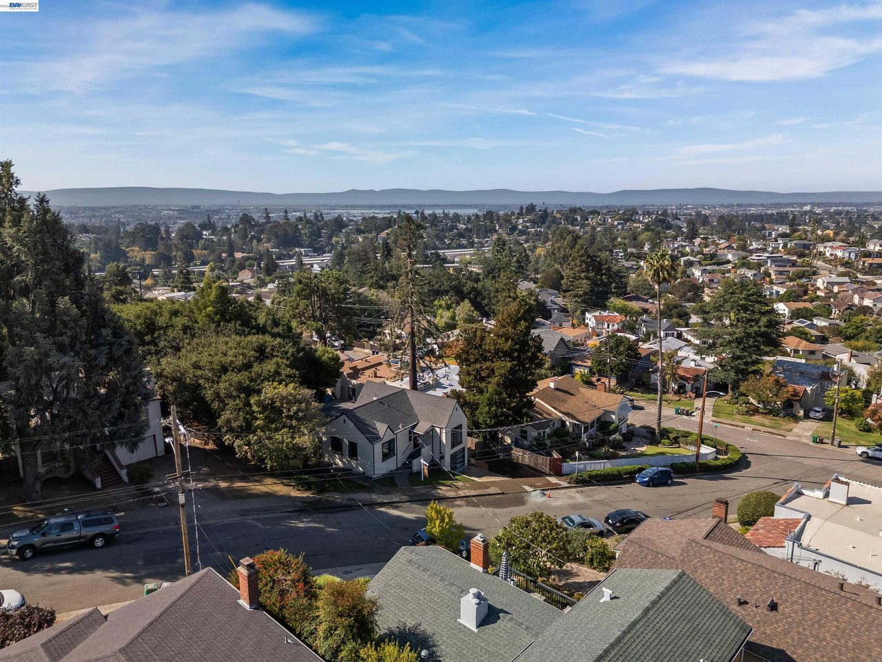 Aerial view over Oakland rooftops and tree-lined streets toward distant water and hills.
