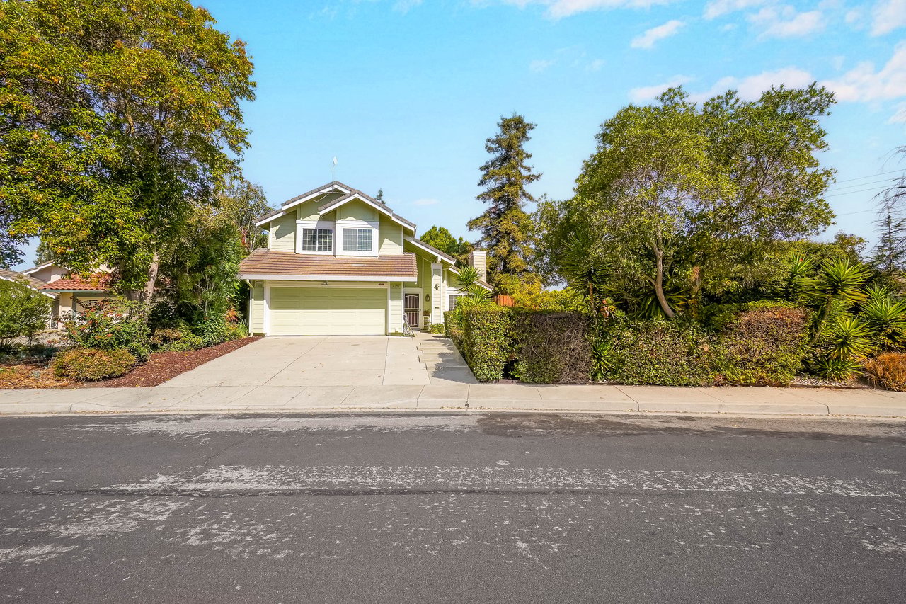Two-story light green house with a double garage, wide driveway, and mature landscaping.