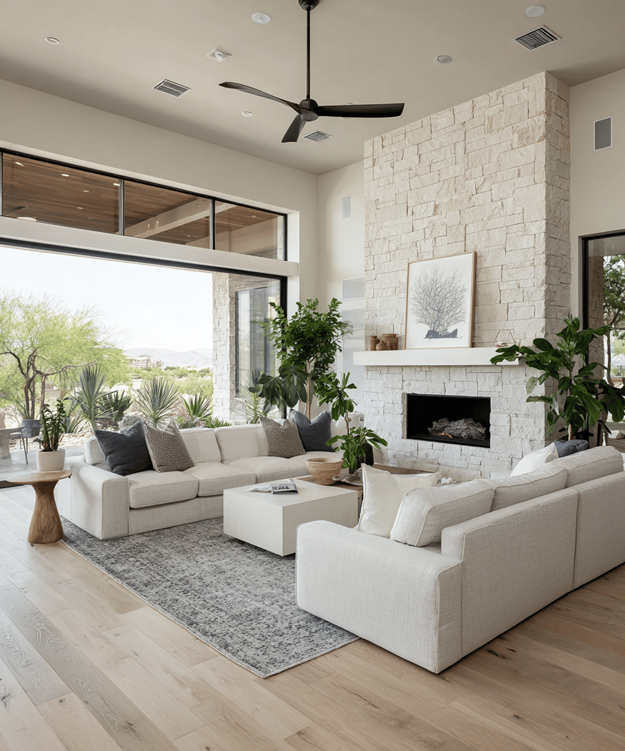 Living room with pale sofas, a tall stone fireplace, and large windows overlooking desert landscaping.