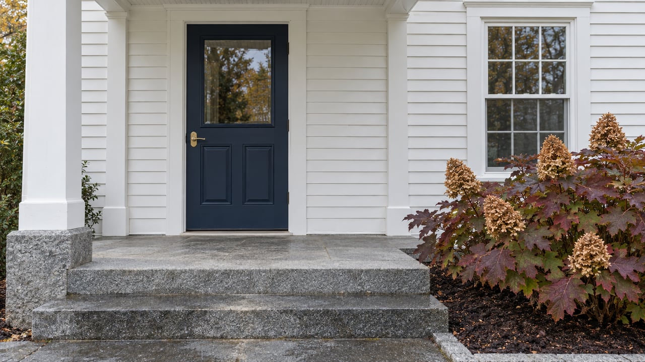 White clapboard home with a navy front door, granite porch steps, brass hardware, and burgundy oakleaf hydrangea beside the entry.