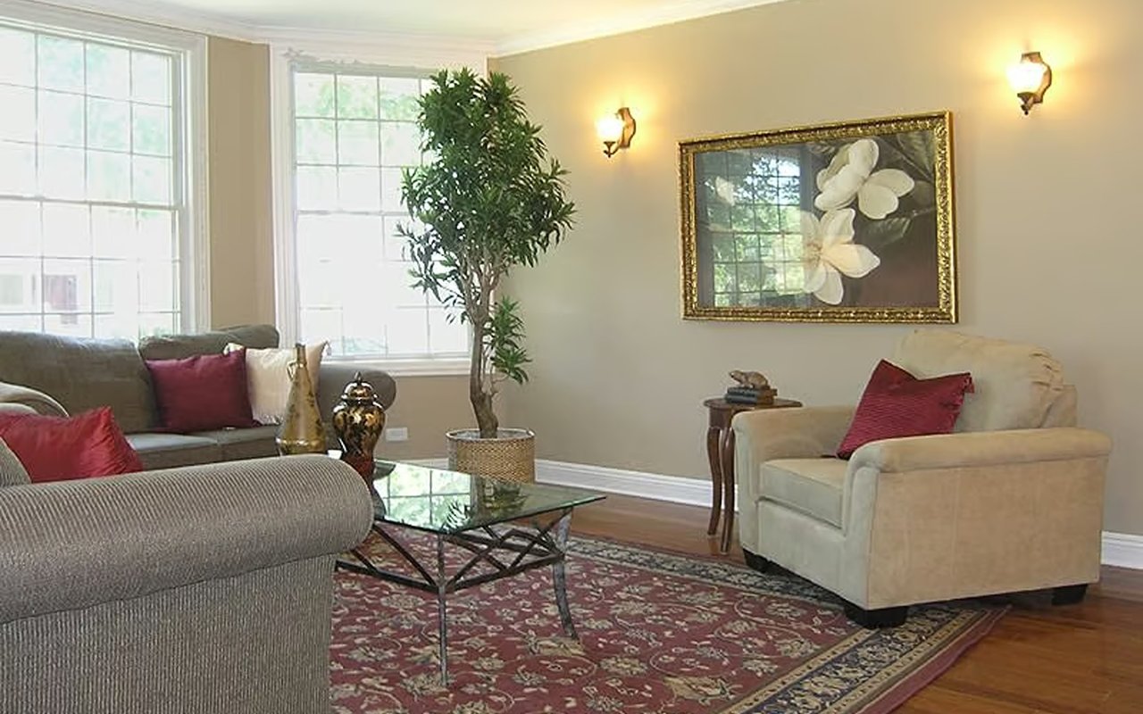 Staged living room with large windows, red accent pillows, a glass coffee table, and patterned rug.