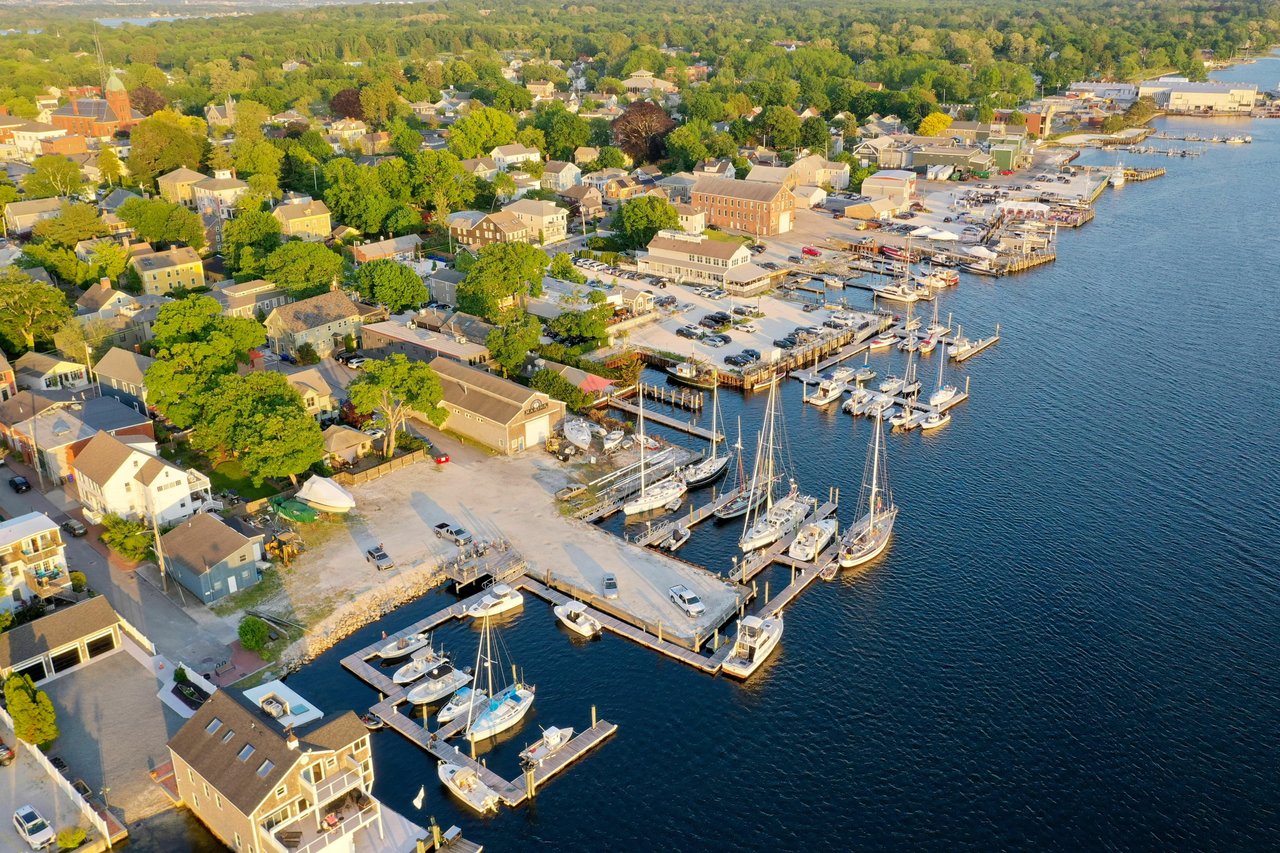 Aerial view of a coastal town and marina, with sailboats docked beside tree-lined streets and waterfront homes.
