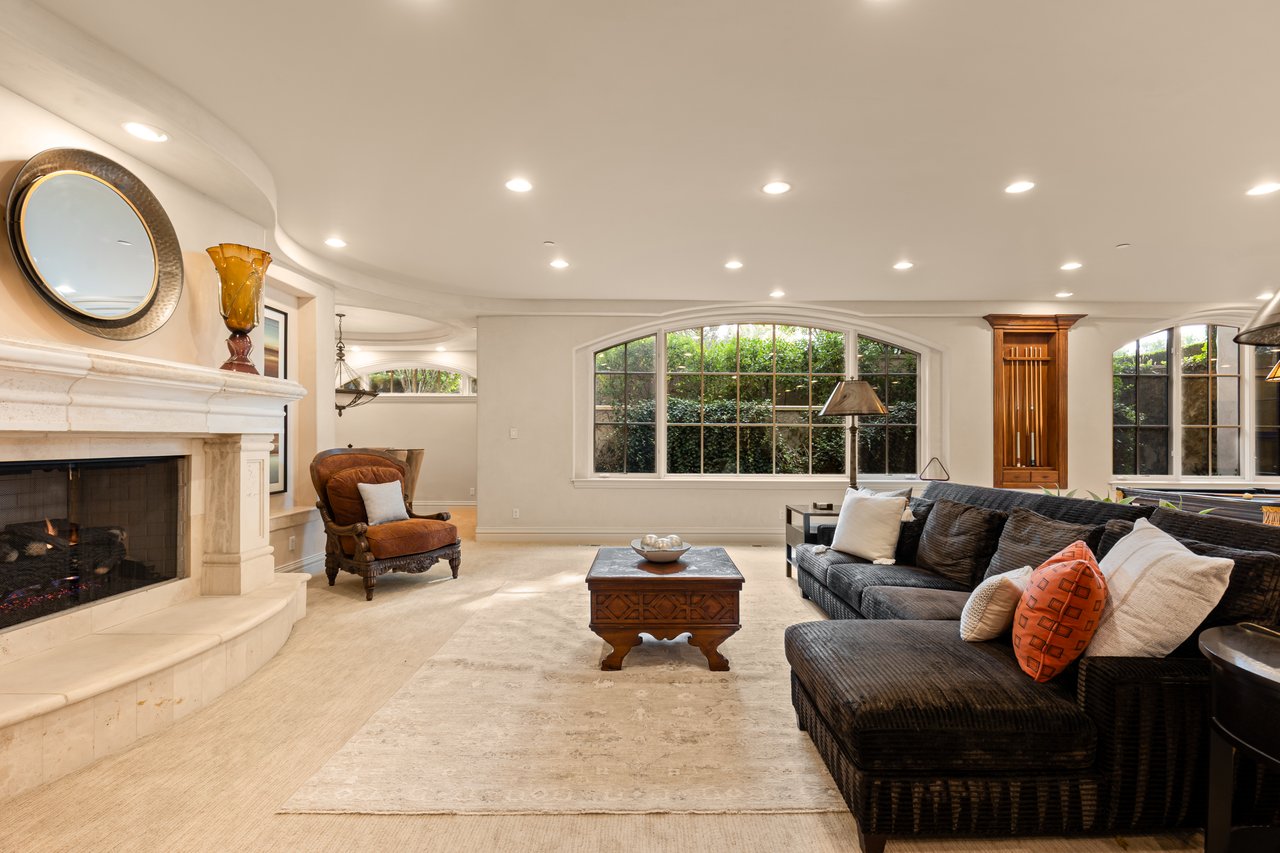 Living room with a curved stone fireplace, dark sectional, and broad arched windows overlooking greenery.