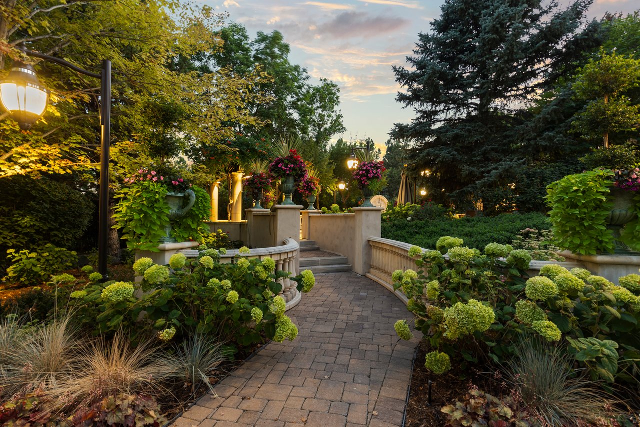 Formal garden path bordered by flowering shrubs and stone balustrades, with lampposts glowing at dusk.