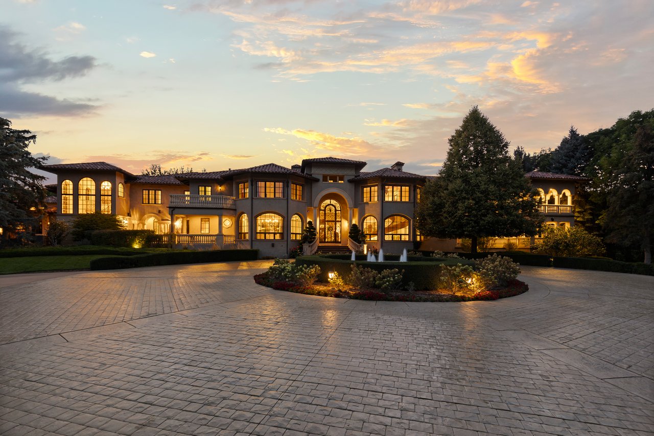 Mediterranean-style estate glowing at dusk, with arched windows, a circular driveway and garden fountains.