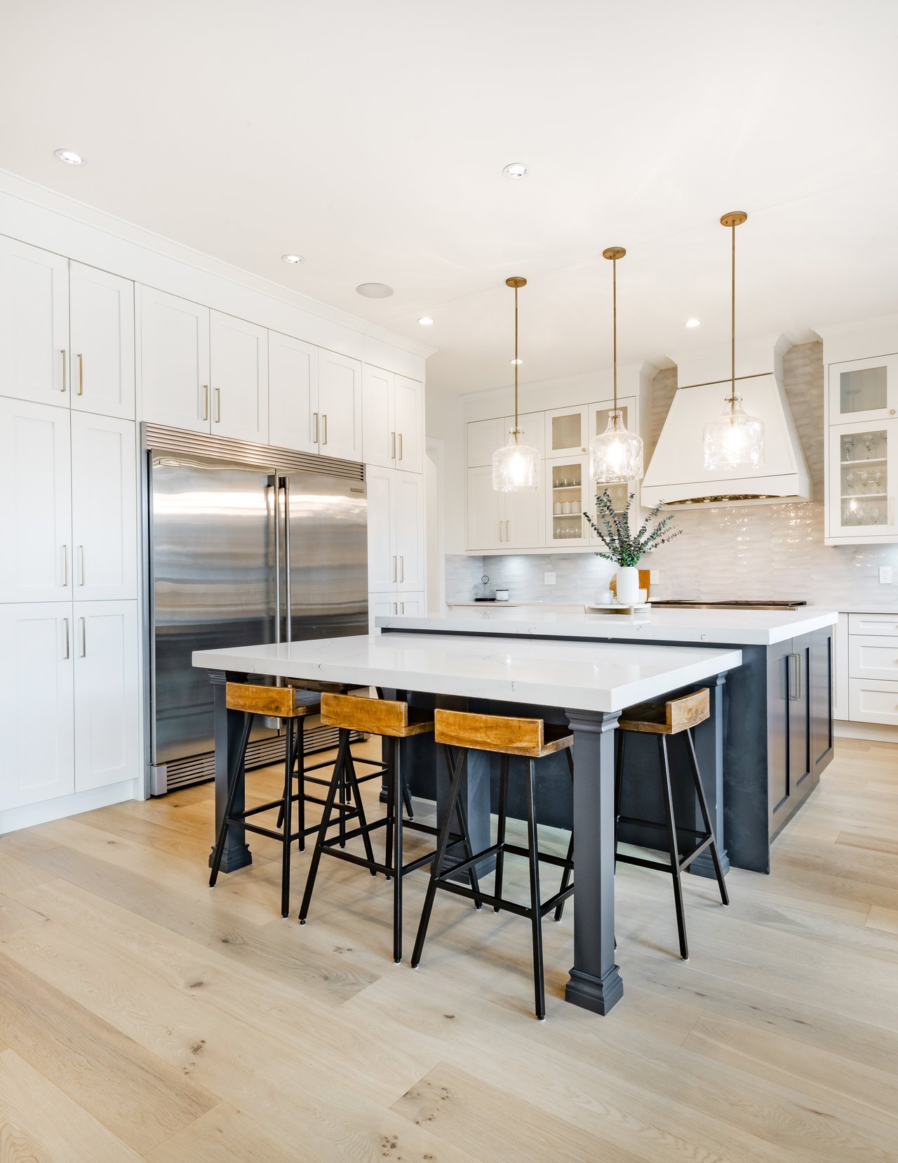 Bright white kitchen with a large island, wooden stools, stainless refrigerator, and pendant lights.
