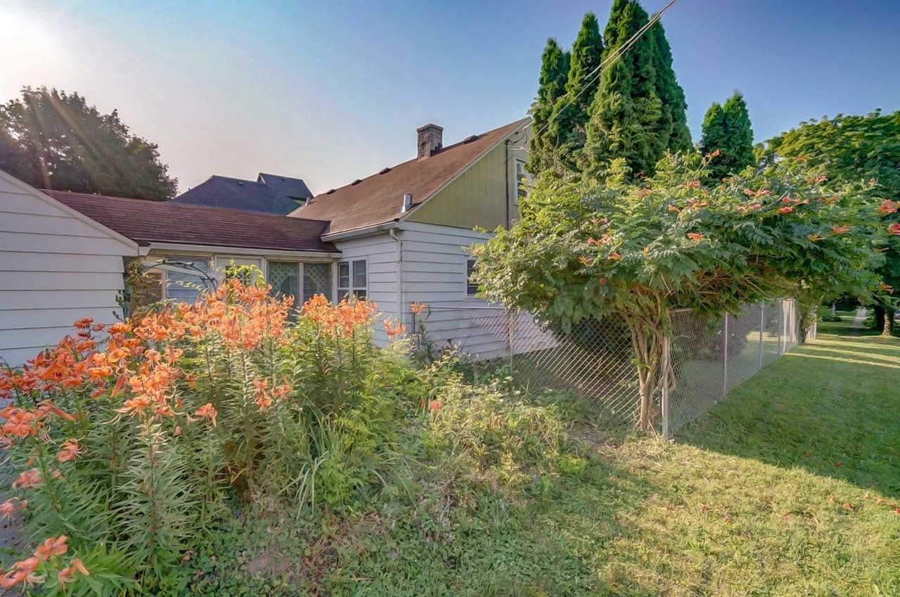 House exterior behind a chain-link fence, with orange flowers and tall evergreens.